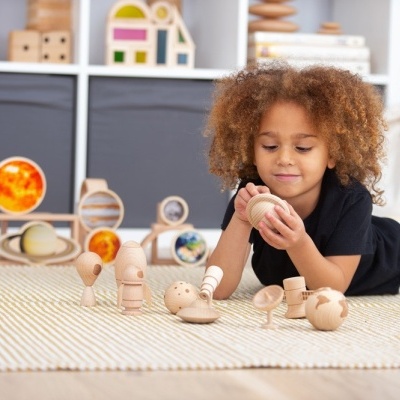Criança brinca com bonecos de madeira sobre tapete em quarto com estantes e brinquedos