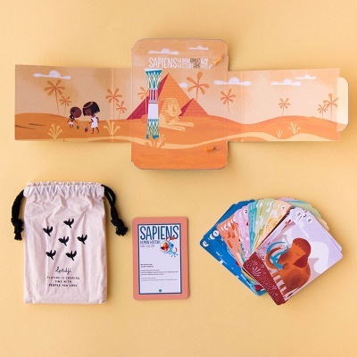 Jogo de cartas SAPIENS com caixa aberta, saco de pano e cartas coloridas sobre fundo amarelo.