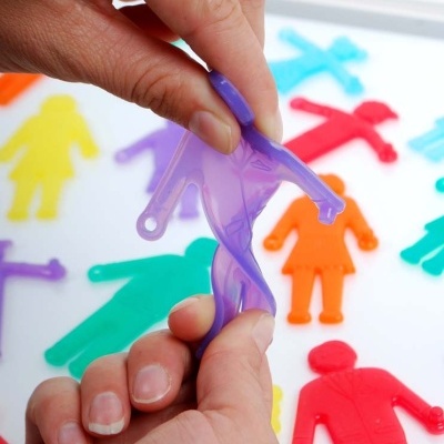 Mãos seguram figura humana de silicone roxa com várias figuras coloridas ao fundo