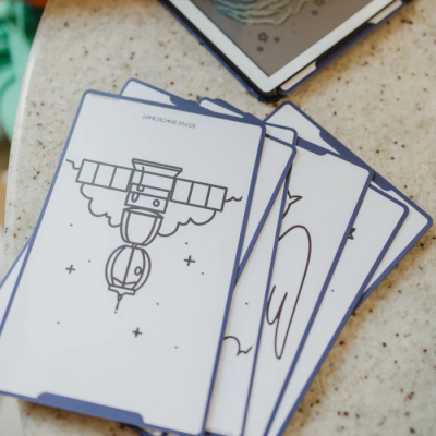 Cartas ilustradas com bordas azuis e desenhos a preto e branco numa superfície clara com tablet ao fundo