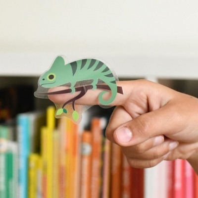 Dedo humano a segurar um autocolante de camaleão verde com riscas pretas em frente a uma estante de livros coloridos