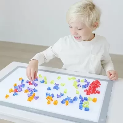 Criança loira com camisola branca a brincar com pequenos objetos coloridos numa base branca num tabuleiro cinzento