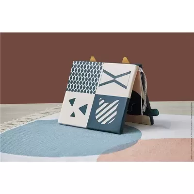 Livro infantil sensorial com capas em tecido com padrões geométricos e alças sobre tapete colorido.