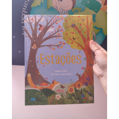 Livro infantil 'Estações' com ilustrações de árvores e animais, segurado por uma mão frente a um fundo colorido