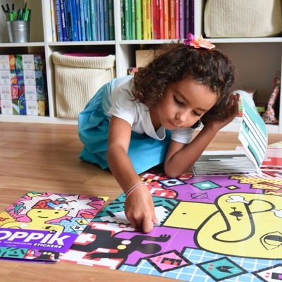 Criança a montar puzzle colorido no chão com caixa 'OPPIK STICKER' ao lado e estantes com organizadores ao fundo