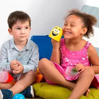 Duas crianças sentadas com almofadas coloridas, menina com marioneta amarela sorridente e chávena com planta