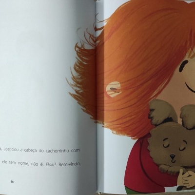 Livro ilustrado aberto com texto em português e desenho de criança ruiva a abraçar cachorro castanho.