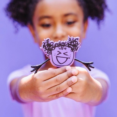 Figura de brinquedo lilás com rosto sorridente e cabelo preto, segurada por uma adolescente com roupa rosa e fundo roxo.