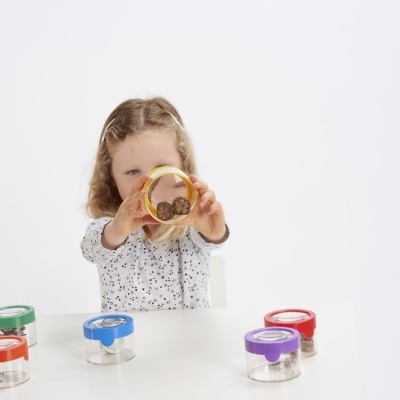 Criança segurando recipiente amarelo com moedas à frente de vários recipientes coloridos numa mesa branca.