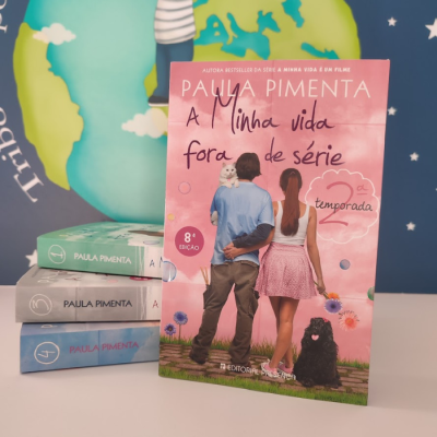 Capa do livro 'A Minha Vida Fora de Série 2ª temporada' de Paula Pimenta com imagem de casal e cão, sobre pilha de livros e fundo mapa do mundo.
