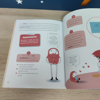 Página de livro ilustrado com texto e personagem vermelho sobre superfície de madeira