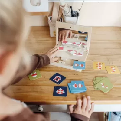 Criança a jogar jogo de cartas com padrões geométricos numa mesa de madeira com espelho e cestos ao fundo