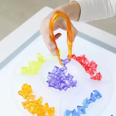 Mão a usar pegador de plástico laranja para apanhar pedaços de plástico colorido em recipiente branco