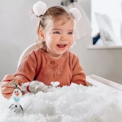 Criança sorridente com camisola rosa claro e pompons brancos no cabelo brinca com neve artificial e bonecos de Frozen na mesa