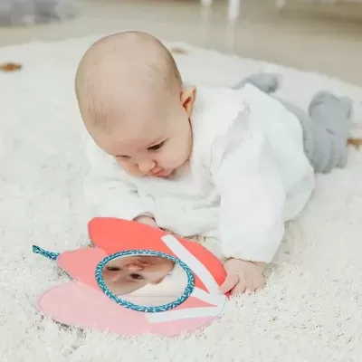 Bebé a olhar para espelho infantil em forma de pétalas rosa e vermelho sobre tapete branco