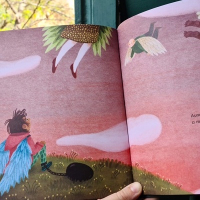 Livro ilustrado aberto com personagem com asas azuis numa paisagem com céu rosa e nuvens.