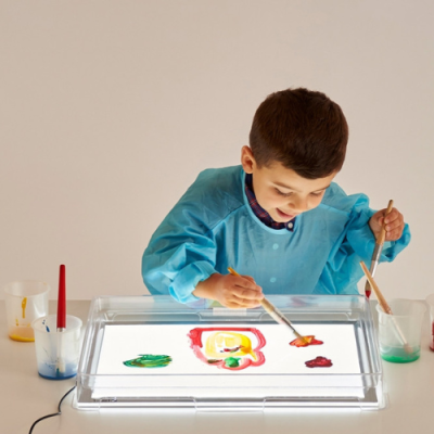 Criança a pintar com pincéis em mesa de luz com tinta colorida e bata azul