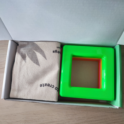 Caixa aberta com saco de tecido e peça plástica quadrada verde e laranja