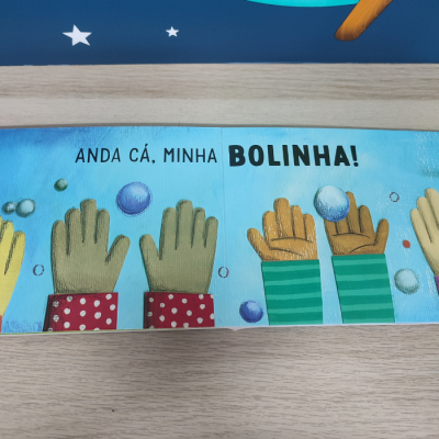 Página de livro infantil com texto em português e ilustrações de mãos a apanhar bolas coloridas.