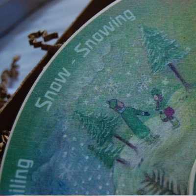 Embalagem redonda com ilustração de pessoas na neve e texto Snow - Snowing