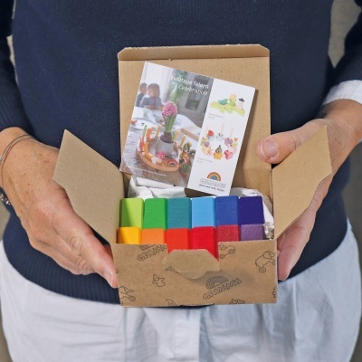 Caixa aberta com blocos de madeira coloridos e folheto dentro, segurada por pessoa com casaco azul e calças brancas.
