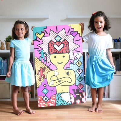 Duas meninas seguram quadro colorido com figura desenhada e elementos gráficos numa sala com estante