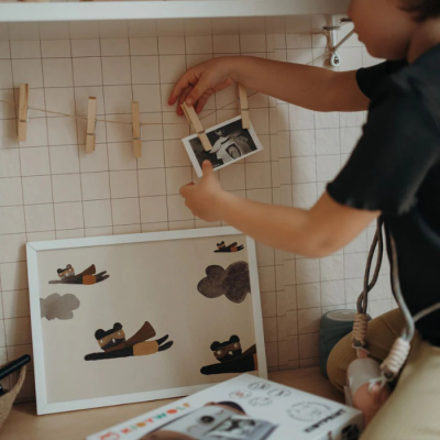 Criança a pendurar foto num pregador de madeira num fio com fundo quadriculado e quadro com desenho de ursos a voar