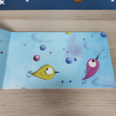 Página de livro infantil com pássaros amarelo e rosa e balões coloridos num fundo azul com nuvens