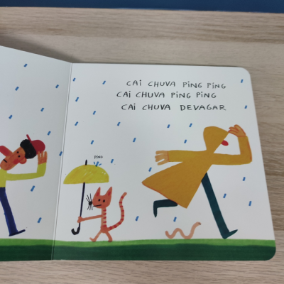 Página de livro infantil com crianças e gato na chuva e texto em português sobre gotas de chuva