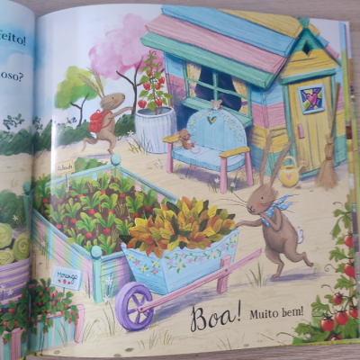 Ilustração colorida de coelhos numa horta com casa de madeira e texto 'Boa! Muito bem!' em livro infantil