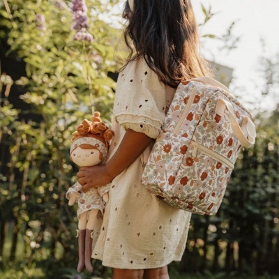 Criança com vestido bege e mochila floral, segurando boneca de pano em ambiente exterior