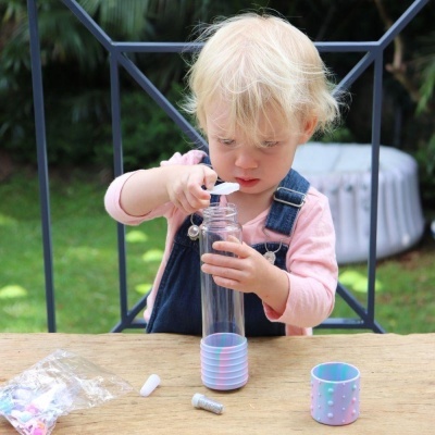 Criança loira mexendo em garrafa transparente com base pastel sobre mesa de madeira ao ar livre