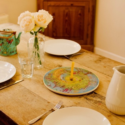 Mesa rústica posta com pratos brancos, copo, jarro, bule verde decorativo, prato colorido com castiçal amarelo e vaso com flores claras