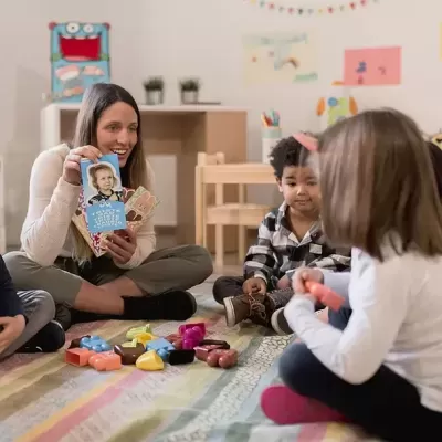 Mulher e crianças sentados no chão com cartão azul e brinquedos coloridos numa sala clara com mobília clara e decoração simples.