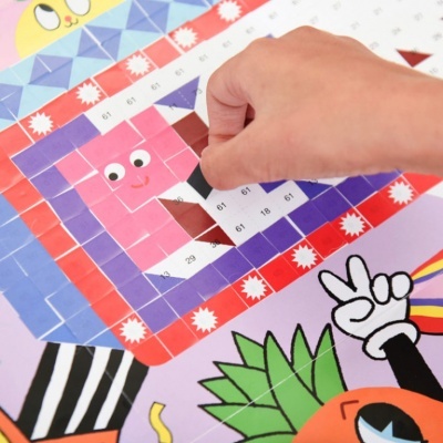 Mão aplicando pedaços coloridos num mosaico de papel com desenhos animados e números.