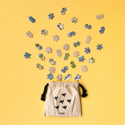 Saco de pano bege com cordões pretos e peças de puzzle coloridas sobre fundo amarelo.