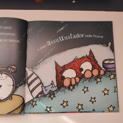 Livro infantil aberto com ilustração de duas personagens na cama e texto em português.