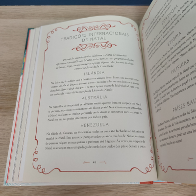 Página de livro em português sobre tradições de Natal, decorada com ornamentos vermelhos, sobre mesa de madeira clara.