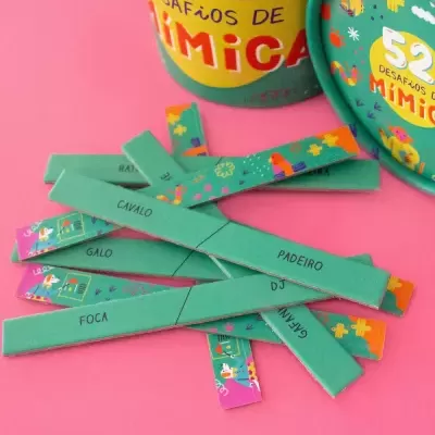 Jogo de tiras de cartão verde com palavras e ilustrações coloridas junto a um recipiente cilíndrico com texto '52 DESAFIOS DE MÍMICA' sobre fundo amarelo e verde