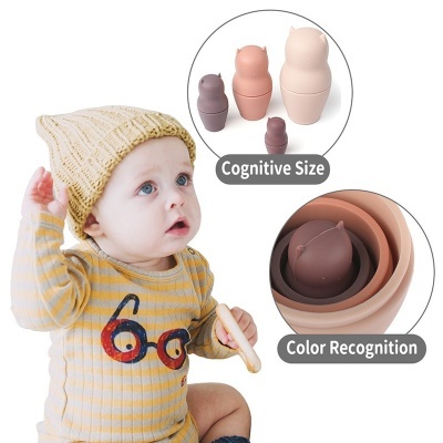 Bebé com gorro e body amarelo segura brinquedo, perto de conjunto de brinquedos empilháveis castanhos e bege