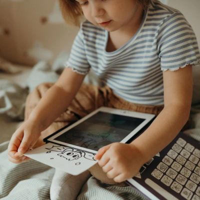 Criança com camisola riscada segurando papel com desenho sentado na cama com tablet e livro