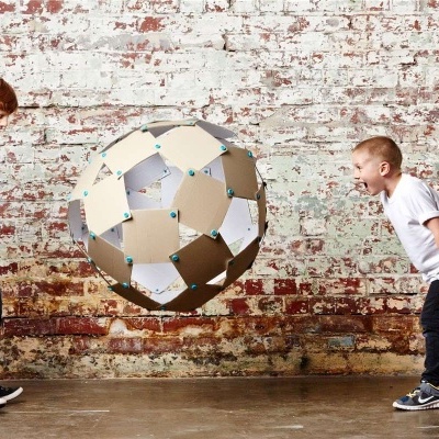 Bola grande feita de cartão entre duas crianças brincando em frente a uma parede de tijolos.