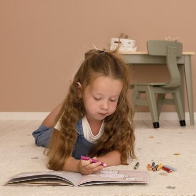 Criança a colorir num livro com lápis de cor num tapete creme com mesa e cadeira ao fundo