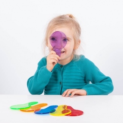 Menina loira com camisola azul turquesa segurando forma plástica roxa circular sobre o olho, com formas plásticas coloridas na mesa branca.