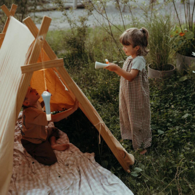 Duas crianças junto a uma tenda de tecido bege em ambiente exterior com candeeiros de mão brancos.