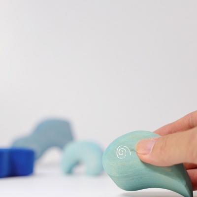 Mão segurando brinquedo de madeira azul claro com desenho branco em espiral e brinquedos desfocados ao fundo.