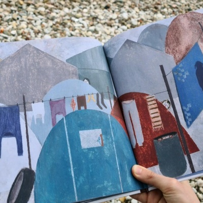 Livro ilustrado aberto com cenas de praia coloridas e mãos a segurá-lo.