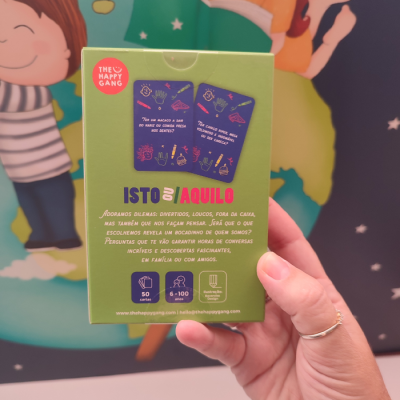 Embalagem verde do jogo ISTO OU AQUILO com texto e ilustrações, segurada por mão com anel