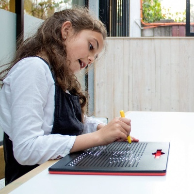 Criança a escrever num quadro braille preto com cruz vermelha numa mesa branca