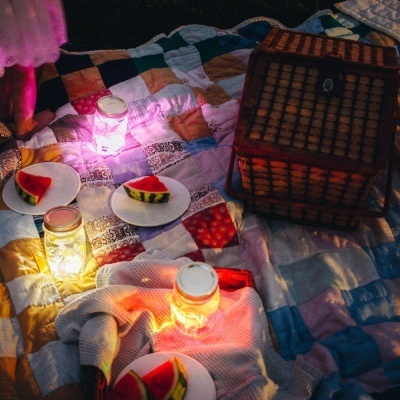 Colcha de retalhos com pratos de melancia, frascos iluminados e cesta de piquenique em ambiente noturno